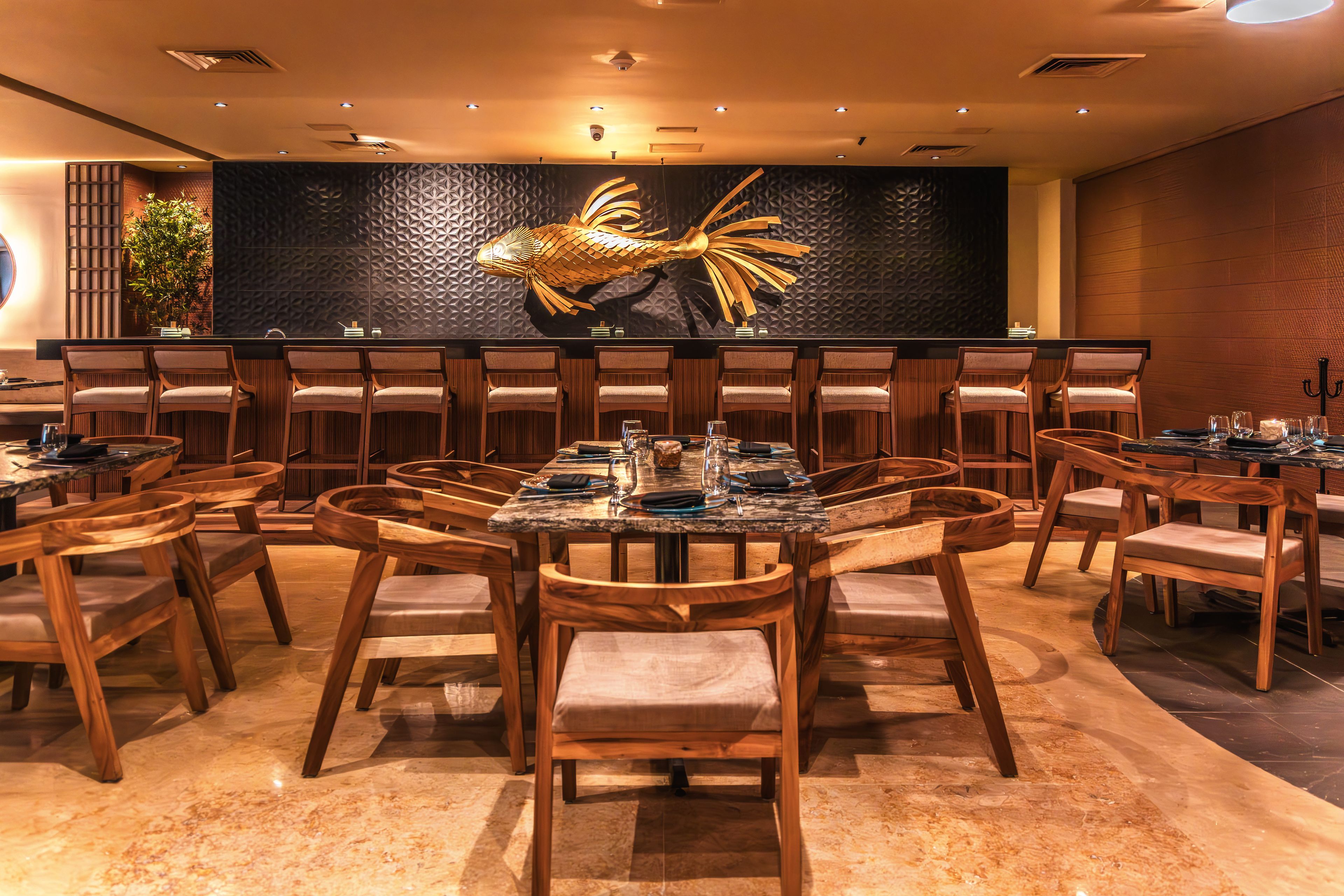 A restaurant with multiple tables and chairs, a decorative wall with a fish sculpture, and a ceiling with recessed lighting.