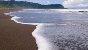 A beach with waves gently lapping onto the shore, a forested hill in the background, and a clear sky. - Canciones del Mar (Jaco)