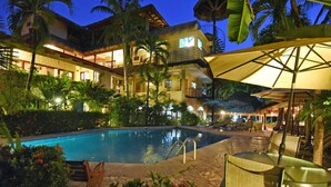 A pool area with lounge chairs and umbrellas, surrounded by lush greenery and a building with multiple balconies. - Canciones del Mar (Jaco)