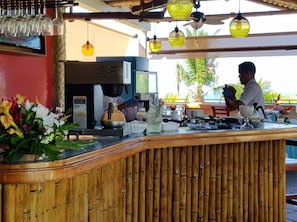 A restaurant counter with a bamboo backdrop, a coffee machine, and a person preparing drinks. - Canciones del Mar (Jaco)