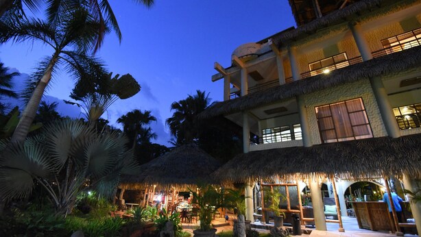 A two-story building with a thatched roof, surrounded by palm trees and a well-lit outdoor area. - Canciones del Mar (Jaco)