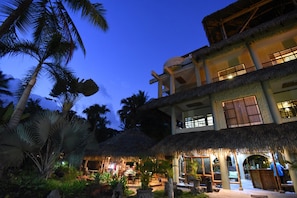 A two-story building with a thatched roof, surrounded by palm trees and a well-lit outdoor area. - Canciones del Mar (Jaco)