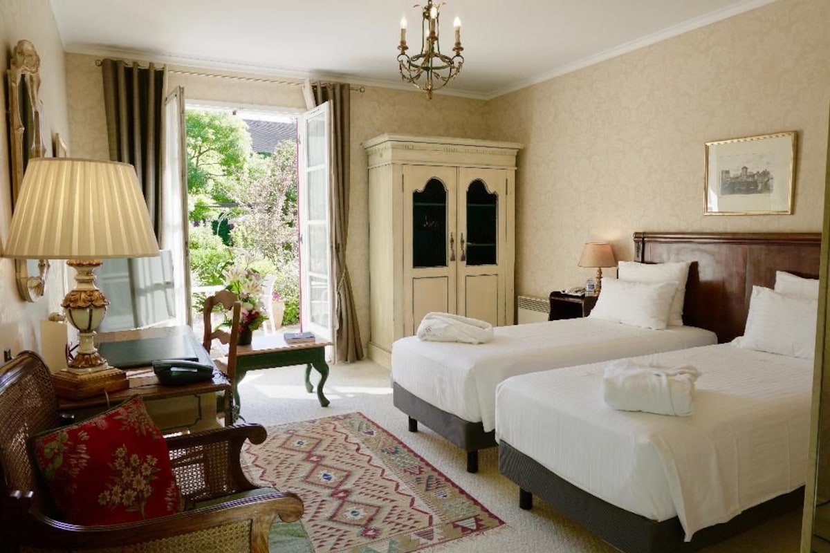 Habitación de hotel con dos camas, un escritorio, una silla, una araña y vistas al exterior.