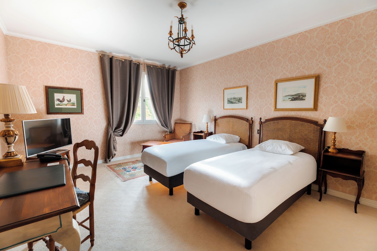 A hotel room with two beds, a desk, a television, and a chandelier.