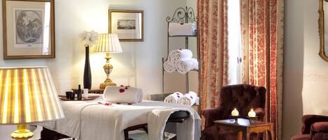 Habitación de hotel con una cama, zona de estar con una silla, una mesa con una vela, una lámpara y una estantería con toallas.