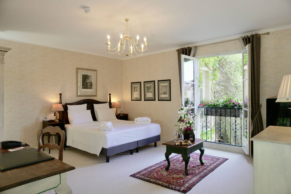 Una habitación de hotel con una cama, mesitas de noche, un escritorio, una silla, un candelabro, cuadros enmarcados y un balcón con plantas.