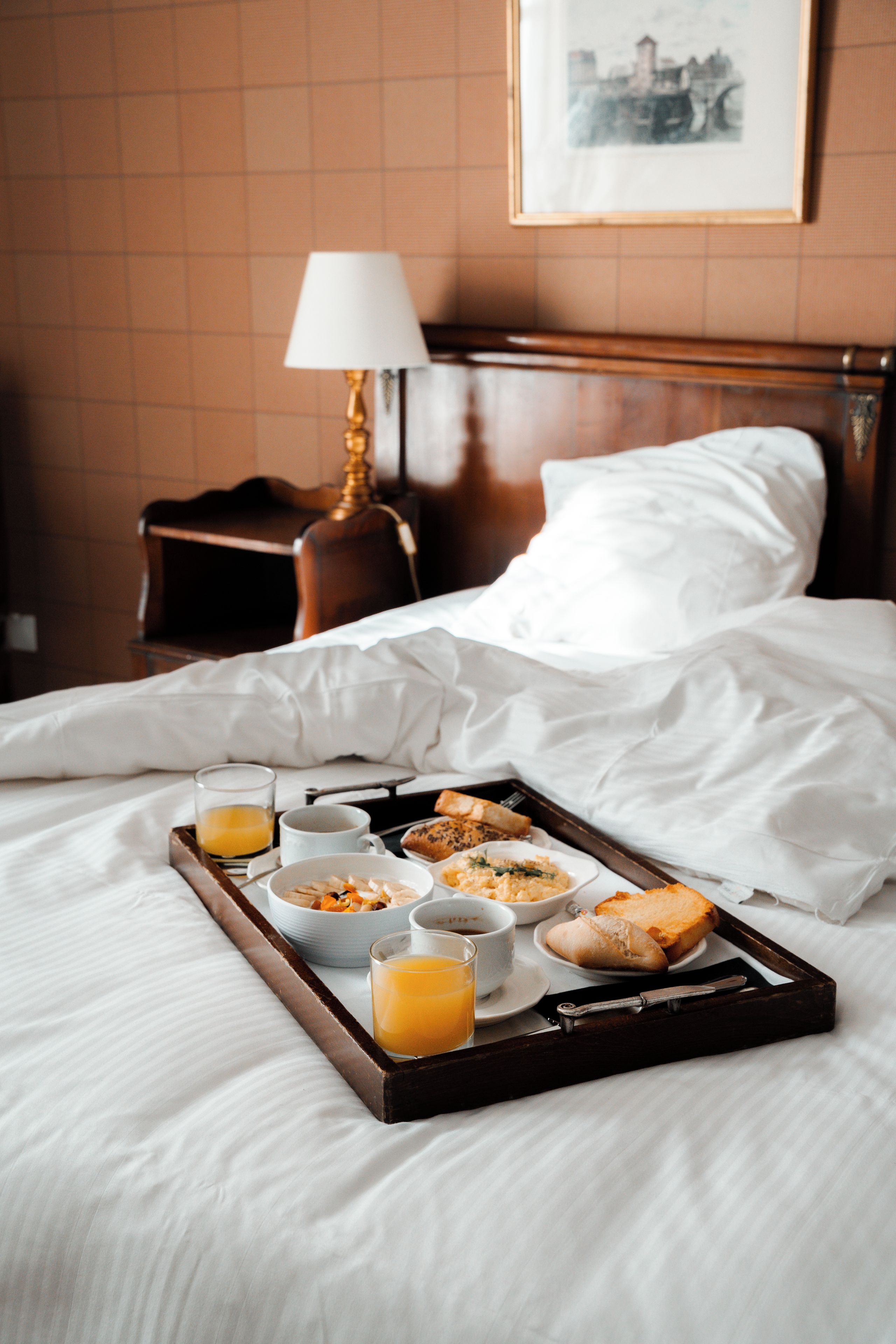 A neatly made bed with white linens, a tray of breakfast including toast, cereal, and juice, and a lamp on a bedside table.