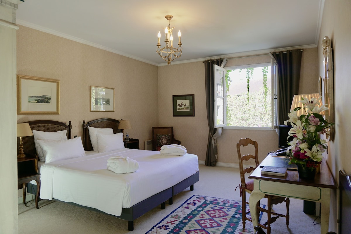 Una habitación de hotel con cama, escritorio, silla, ventana con cortinas y candelabro.