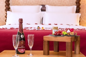 A bed with white linens and rose petals, a bottle of champagne, two glasses, and a small table with fruit and pastries.
