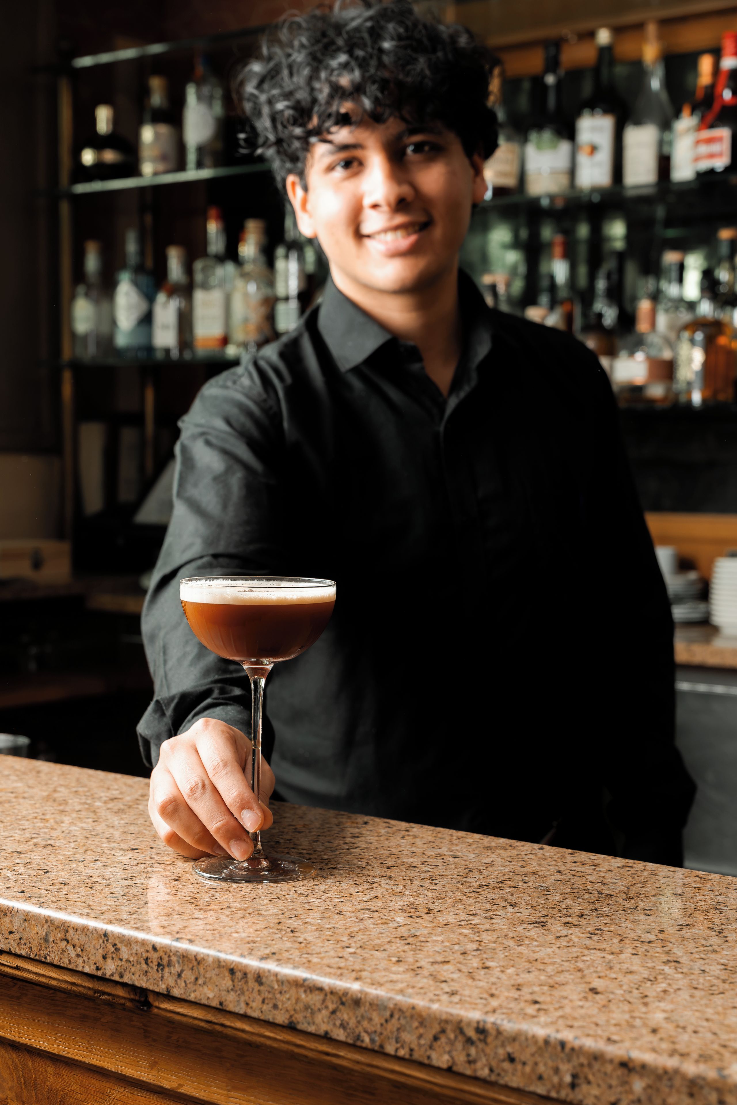 Eine Person hält ein Cocktailglas an der Bar, im Hintergrund sind Regale mit Flaschen zu sehen.