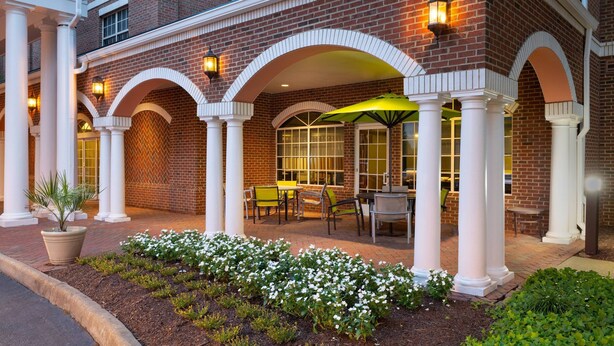 A brick building with white columns and arches, outdoor seating with tables and chairs, and a well-maintained garden with white flowers. - Springhill Suites by Marriott Williamsburg (Williamsburg)