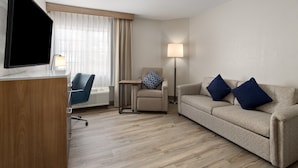 Une chambre d’hôtel avec un bureau en bois, une télévision, un canapé avec deux coussins, une chaise avec un coussin à motifs et un lampadaire.