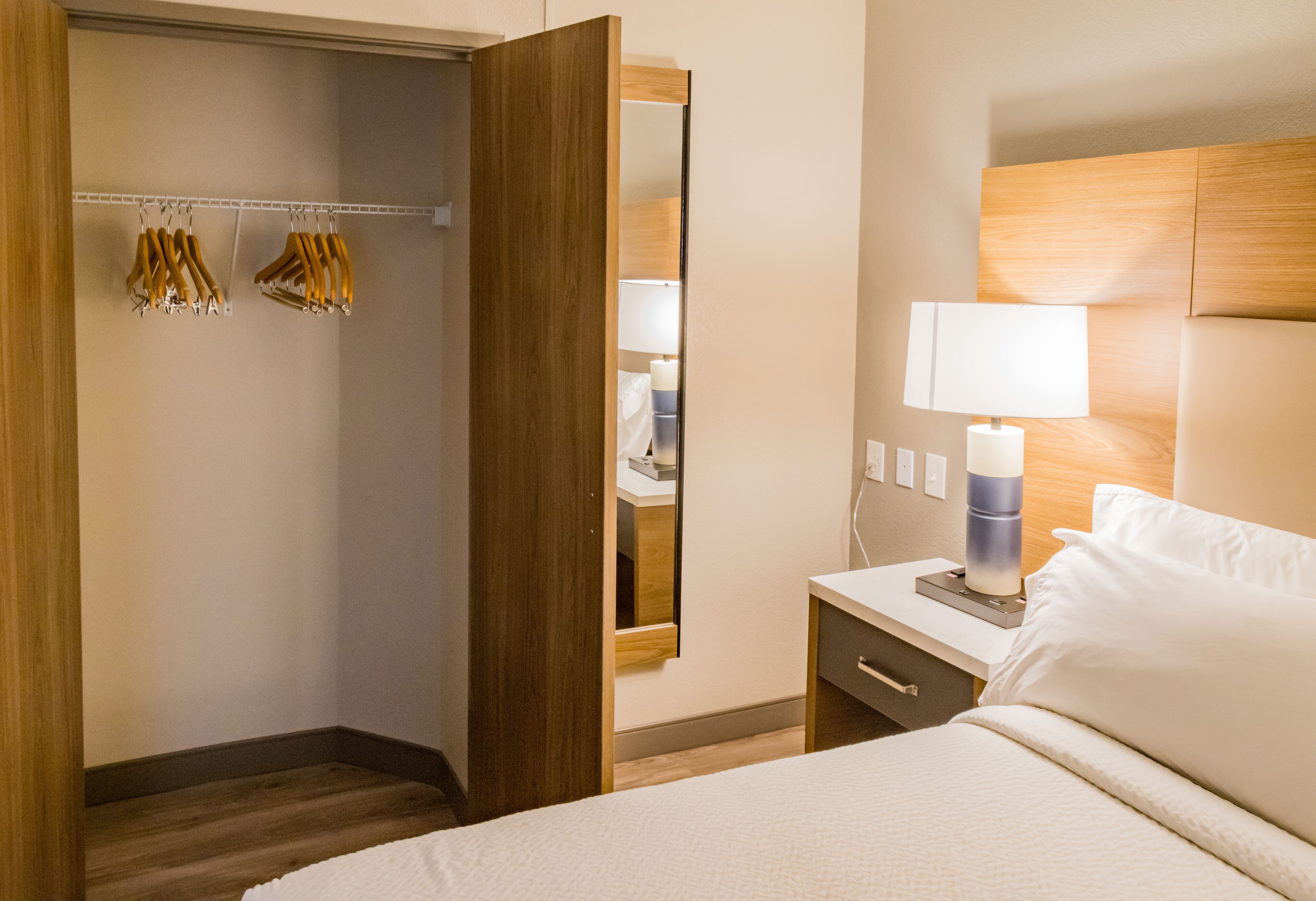 A hotel room with a bed, bedside table, lamp, and a wooden wardrobe with hanging clothes.