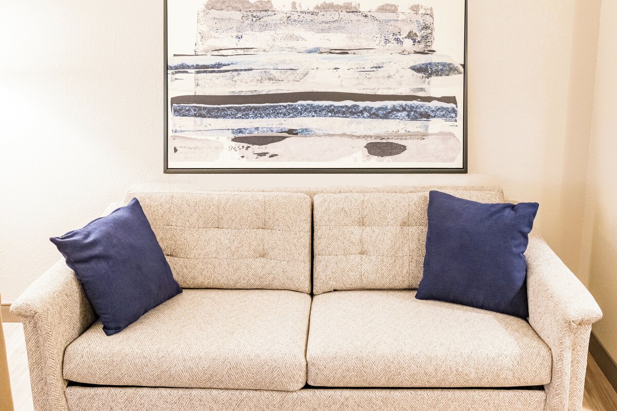 Ein beiges Sofa mit zwei dunkelblauen Kissen, darüber ein gerahmtes abstraktes Gemälde und ein Holzboden.