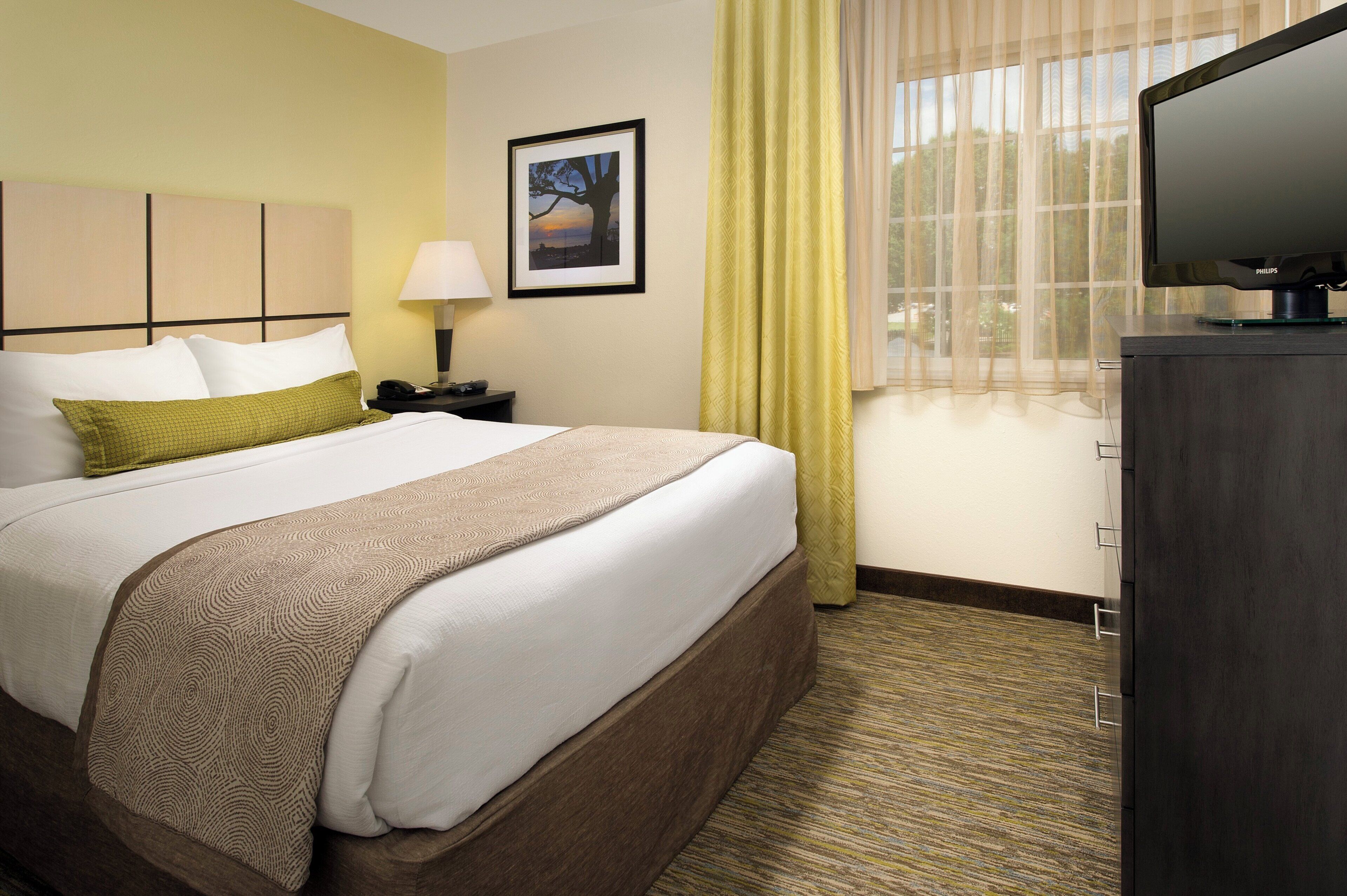 A hotel room with a bed, a nightstand, a lamp, a television, and a window with curtains.