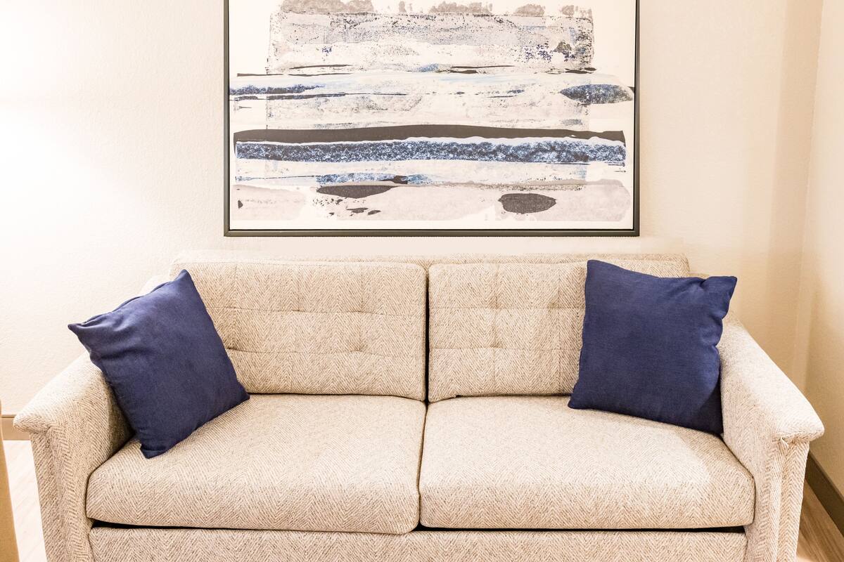 Un canapé beige agrémenté de deux coussins bleu foncé, un tableau abstrait encadré au-dessus, et un plancher en bois.