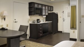 A hotel room with a bed, a desk, a chair, a kitchenette with a microwave, refrigerator, and cabinets, and a bathroom door.