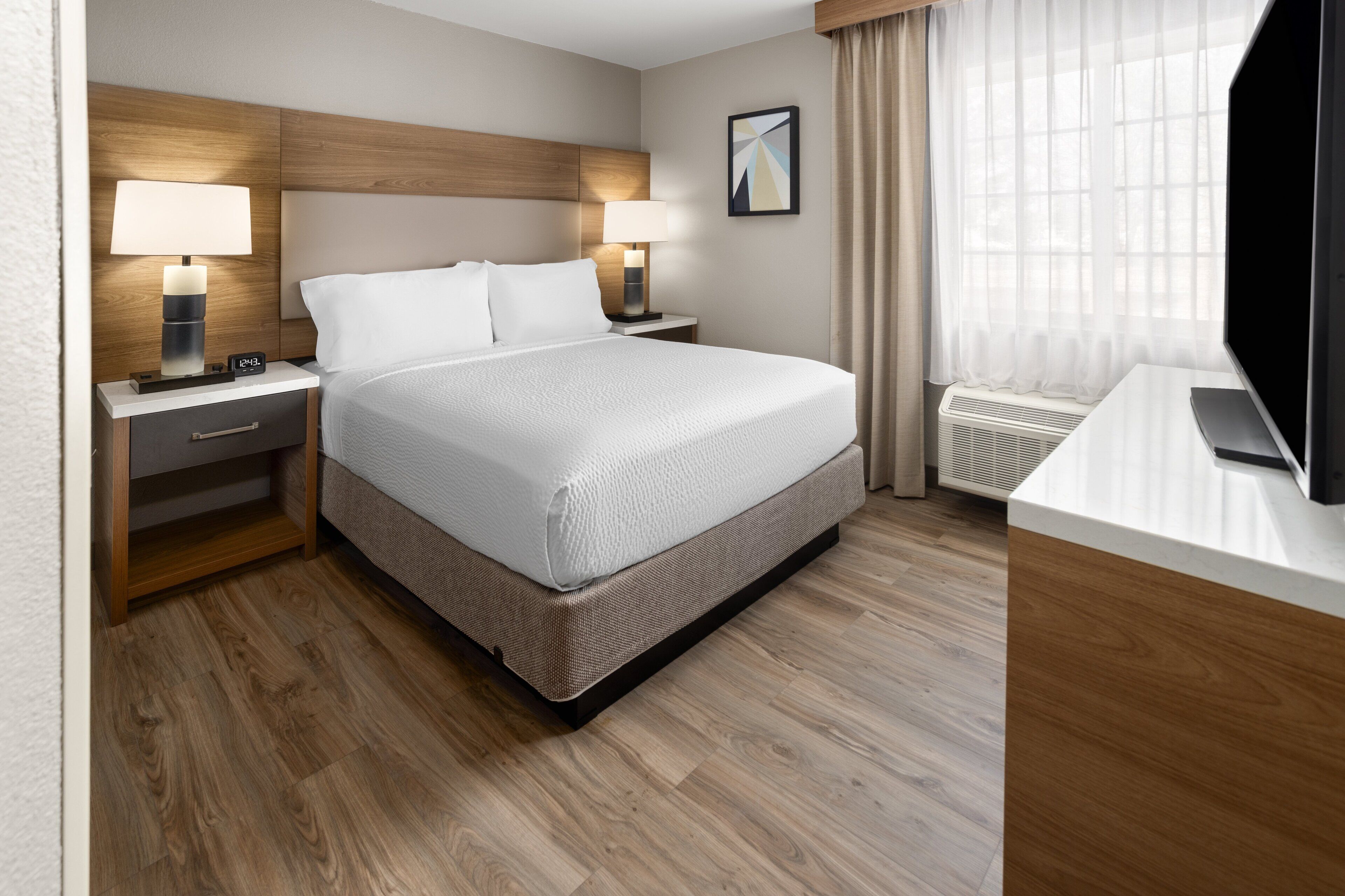 A hotel room with a large bed, two bedside tables with lamps, a flat-screen TV, a desk, and a window with curtains.