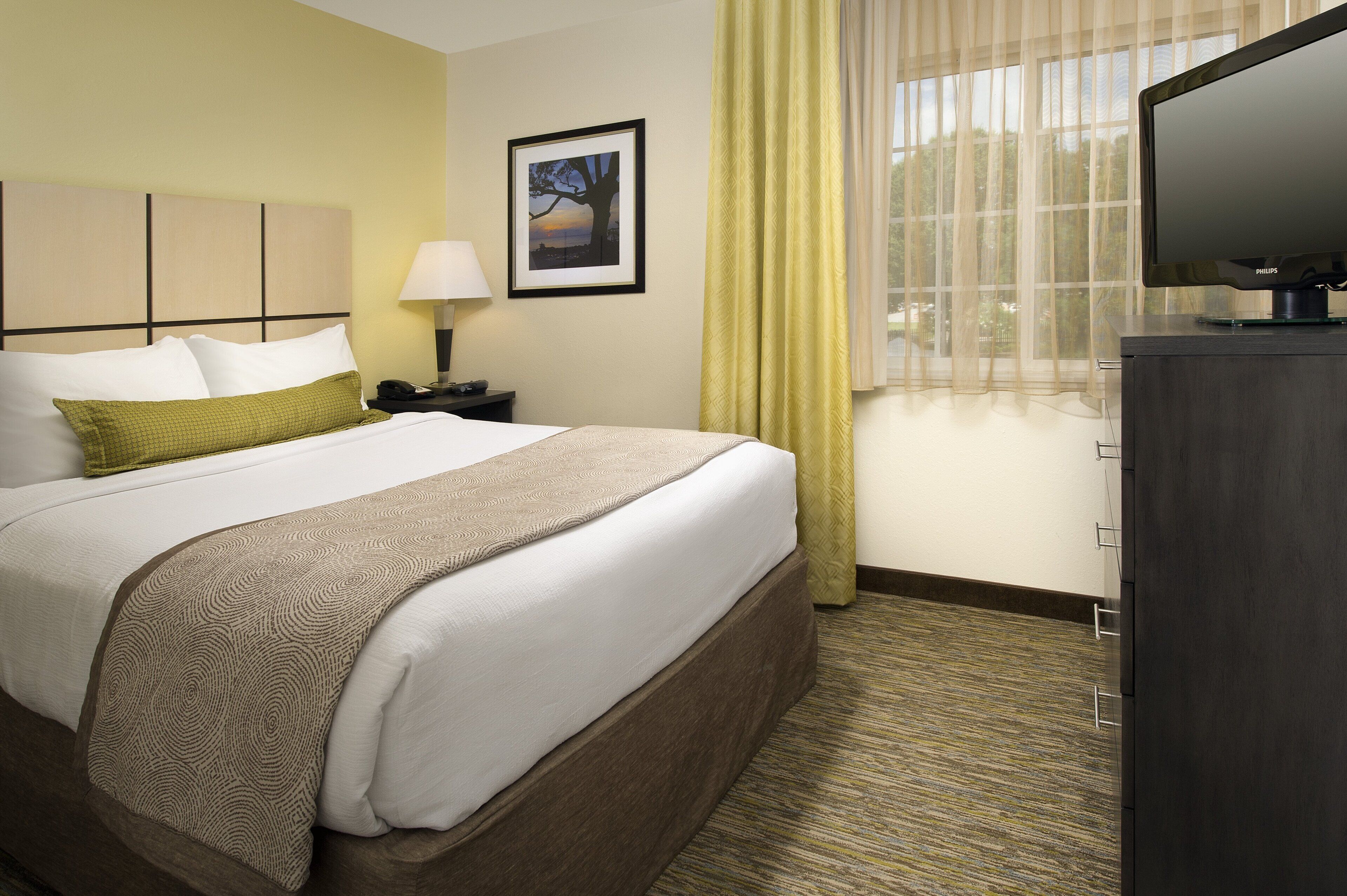 A hotel room with a bed, a nightstand, a lamp, a television, and a window with curtains.