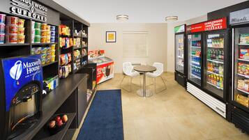 Zona de aperitivos y bebidas con estantes llenos de diversos productos, un refrigerador y un área de descanso con mesa y sillas.