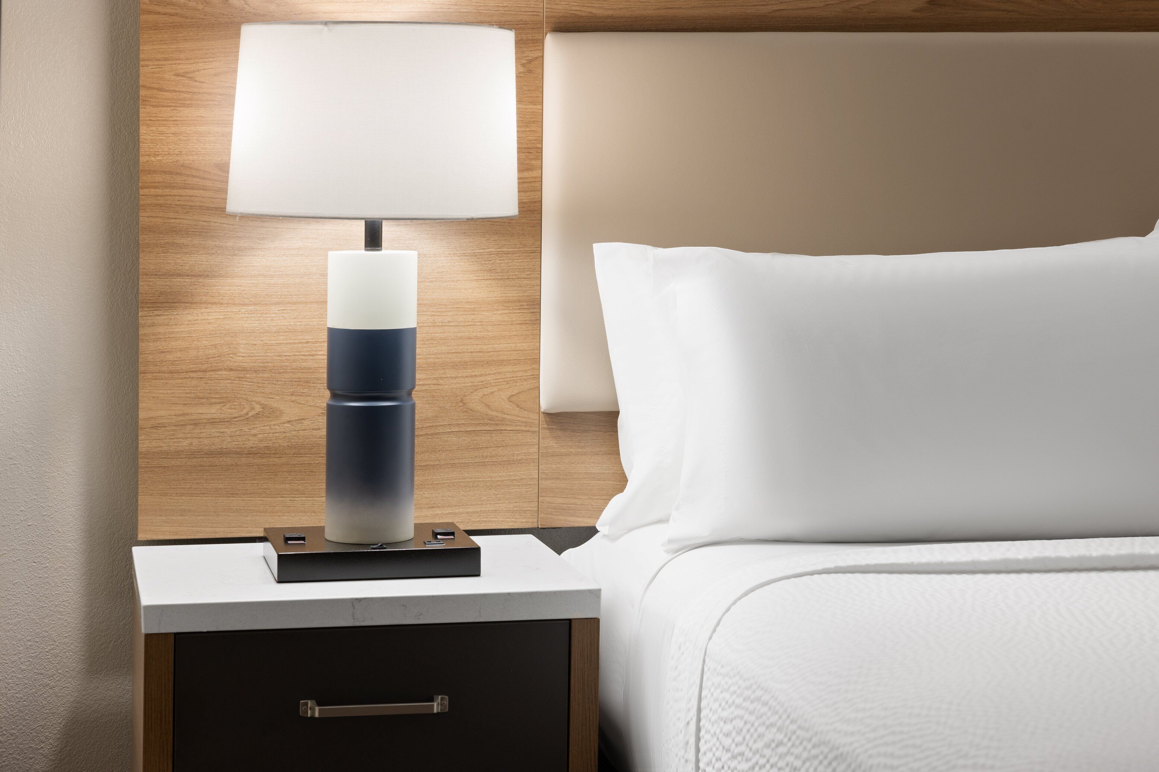 A neatly made bed with white linens, a bedside table with a lamp, and a wooden headboard.