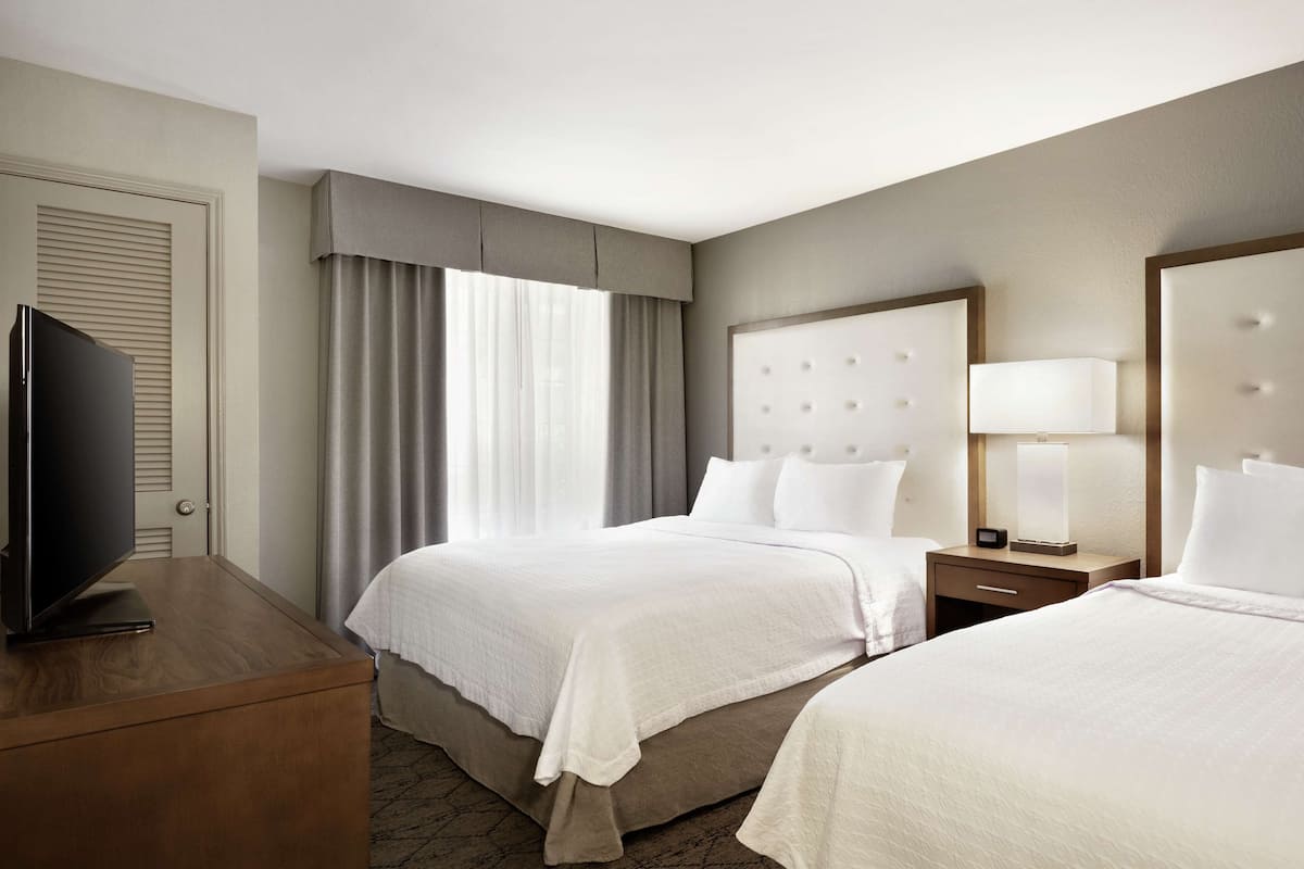 A hotel room with two beds, a television on a wooden dresser, and a nightstand with a lamp.