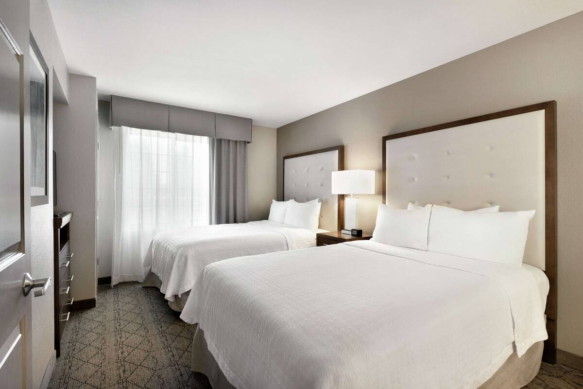 A hotel room with two beds, a large mirror, a bedside table with a lamp, and a window with curtains.