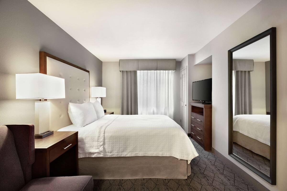 A hotel room with a bed, a desk, a chair, a TV, and a large mirror.