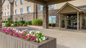 Entrada de hotel con una galería techada, fachada de piedra y un parterre con flores rosas y blancas.