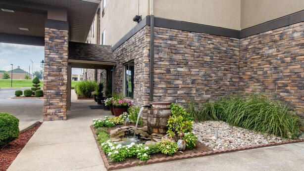 Un edificio con fachada de piedra, un vestíbulo cubierto y un jardín ajardinado con fuente.