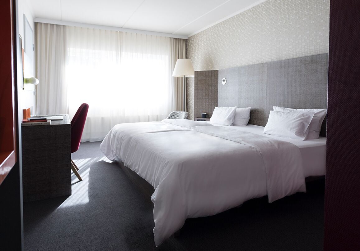 A hotel room with a large bed, a desk with a chair, a lamp, and a view of the outdoors through sheer curtains.