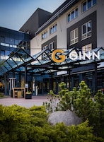 Entrada moderna de hotel com uma cobertura de vidro, uma placa com a inscrição "GOINN" e um elemento decorativo em pedra em primeiro plano.