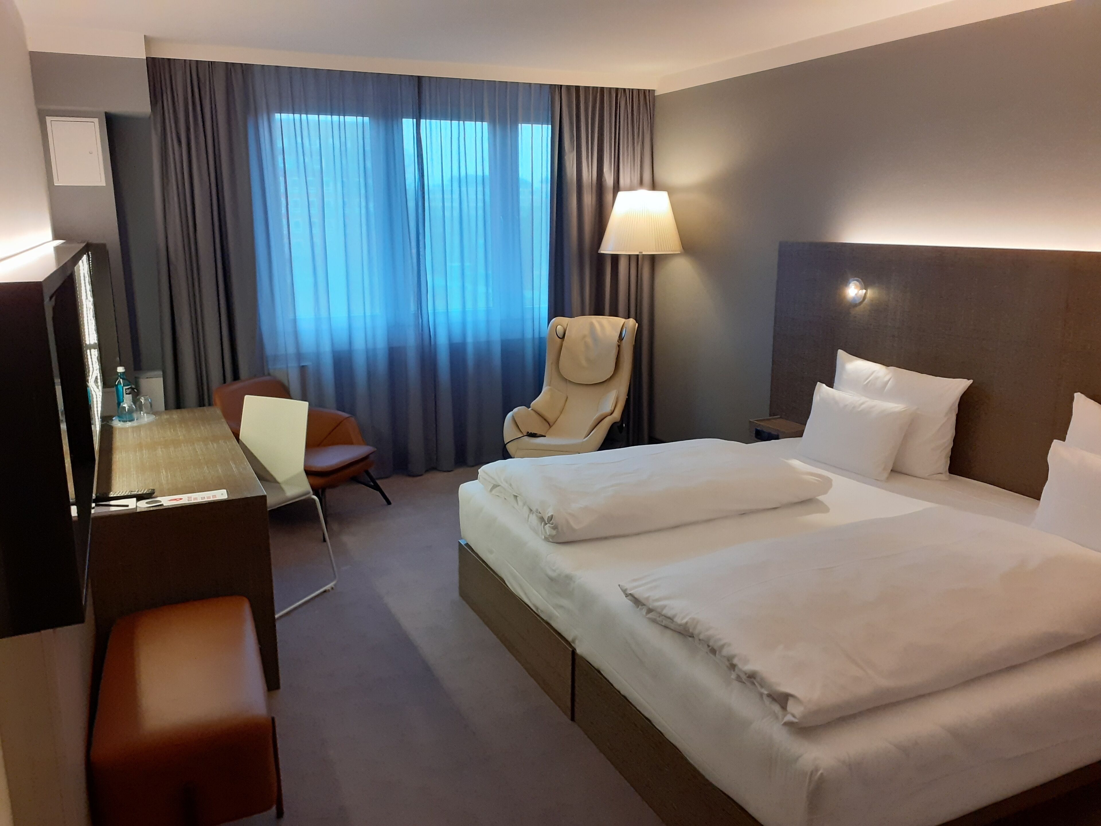 Habitación de hotel con cama, escritorio, silla, lámpara y ventana con cortinas.