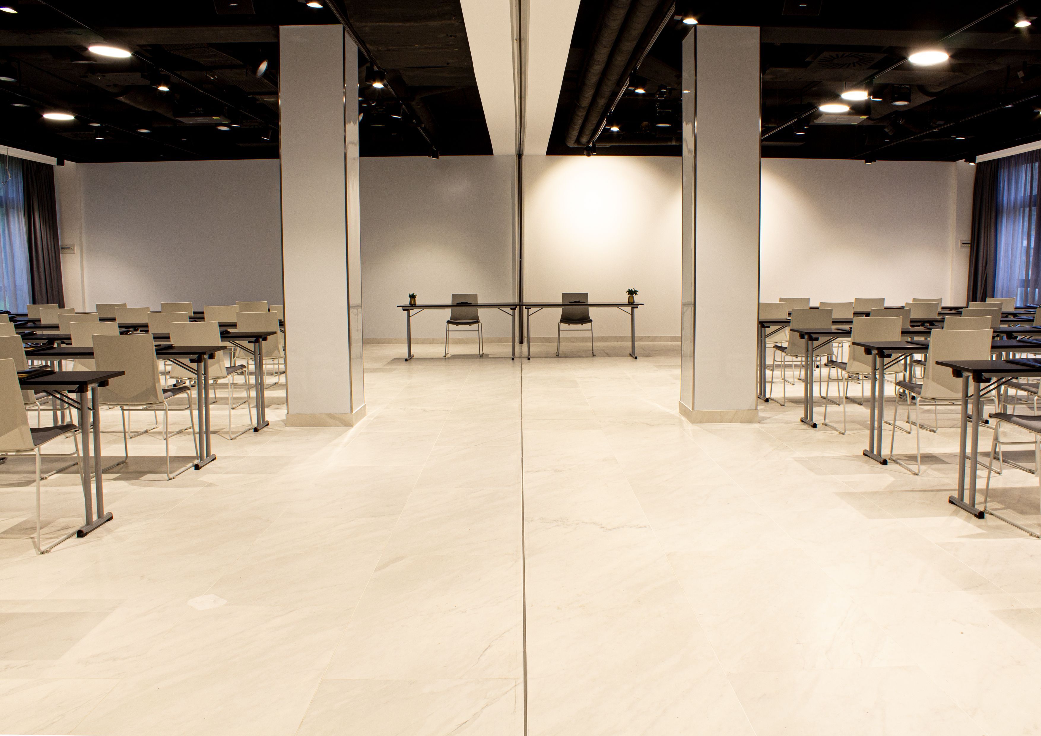 A conference room with a symmetrical layout, featuring rows of tables and chairs, a central aisle, and a plain white wall.