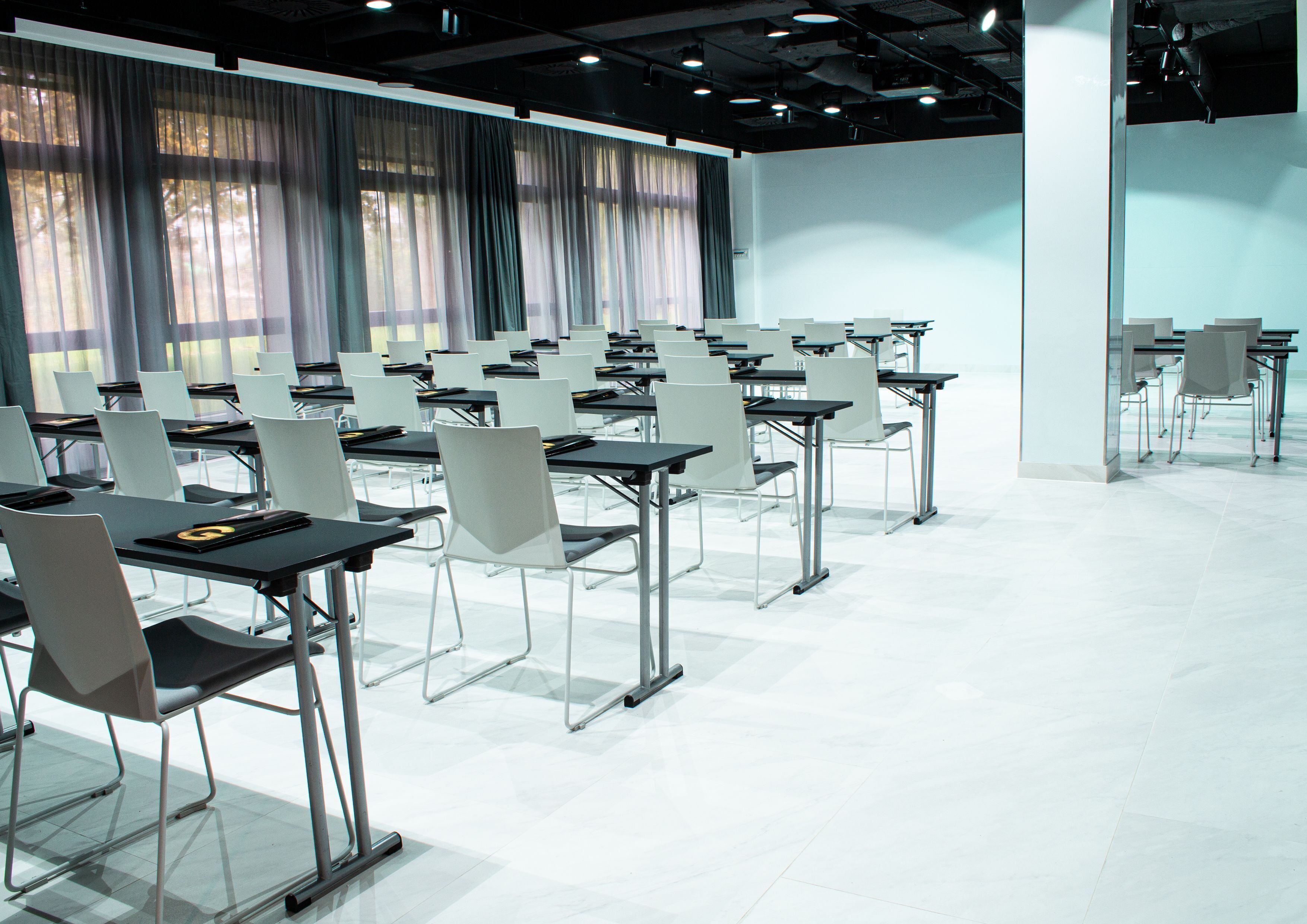 Un salón de conferencias con filas de mesas y sillas, ventanas grandes con cortinas y suelo de baldosas.
