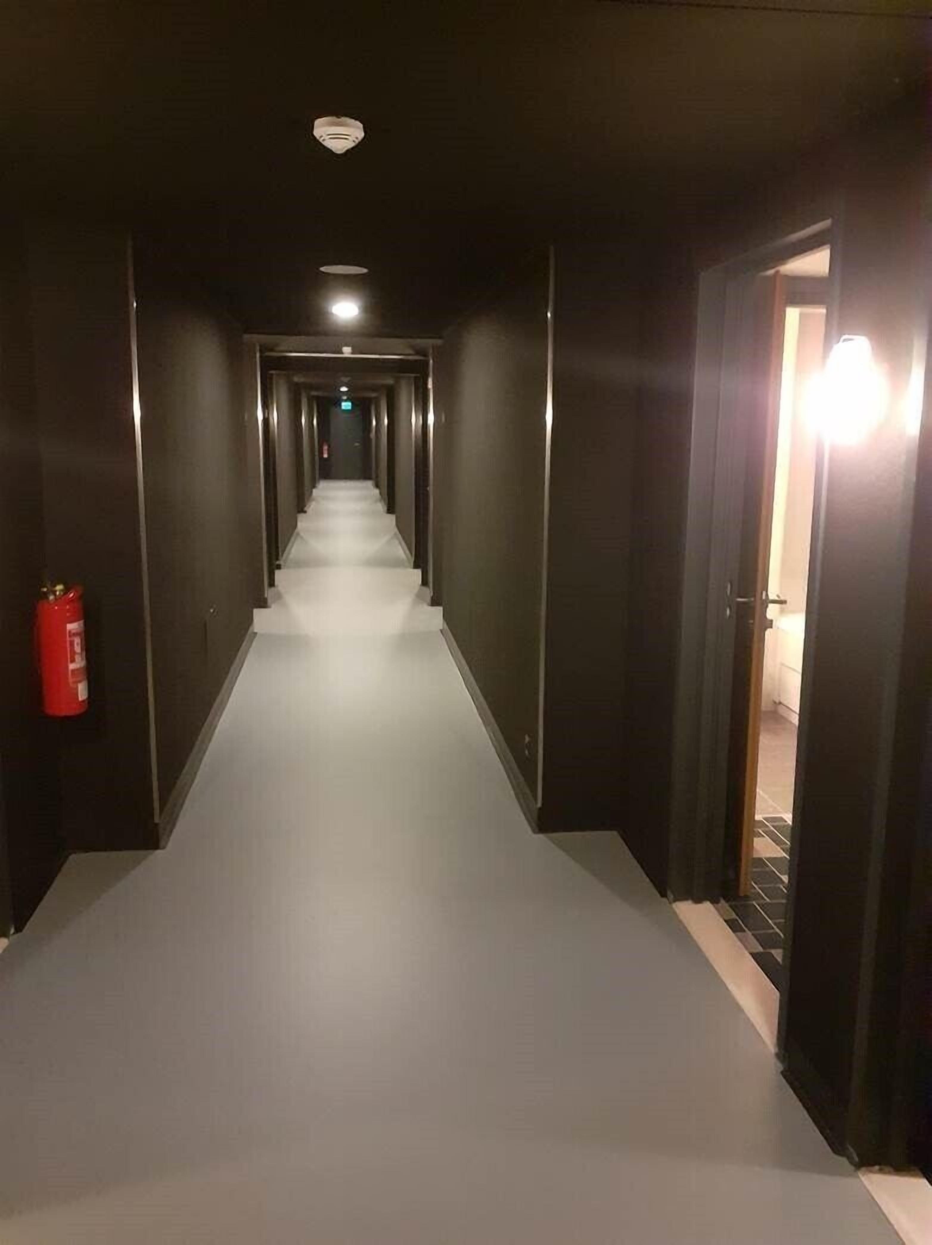 Un long couloir étroit, doté d’un sol recouvert de moquette, bordé de murs sombres et muni d’un extincteur sur le côté gauche.