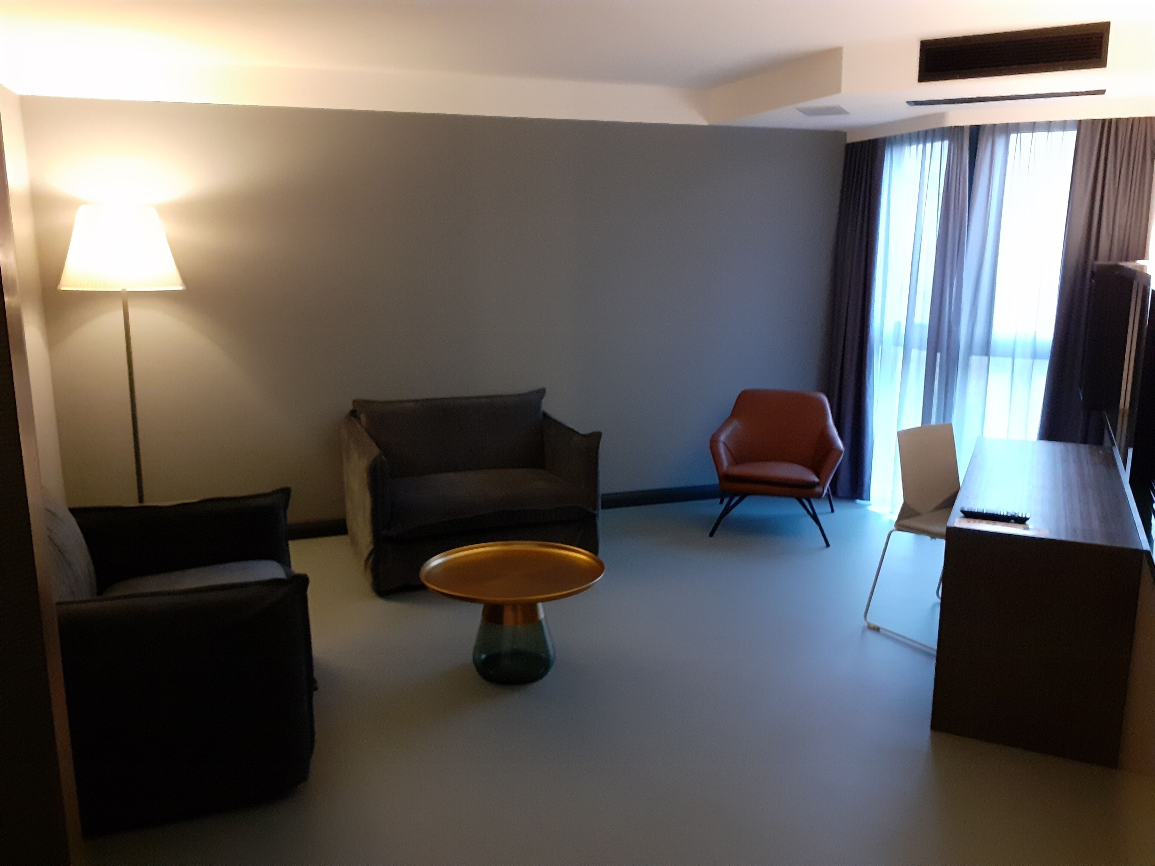 Ein Hotelzimmer mit Sofa, Sessel und einem kleinen Tisch. Eine Lampe, ein Fernseher und ein Fenster mit Vorhängen sind vorhanden.