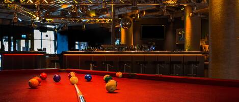 A pool table with balls and a cue stick, a bar area with stools, and a large hanging light fixture.