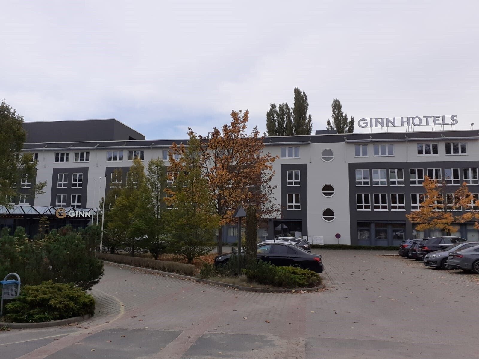 Ein GINN Hotels Gebäude mit Parkplatz und begrüntem Bereich mit Bäumen und Sträuchern.