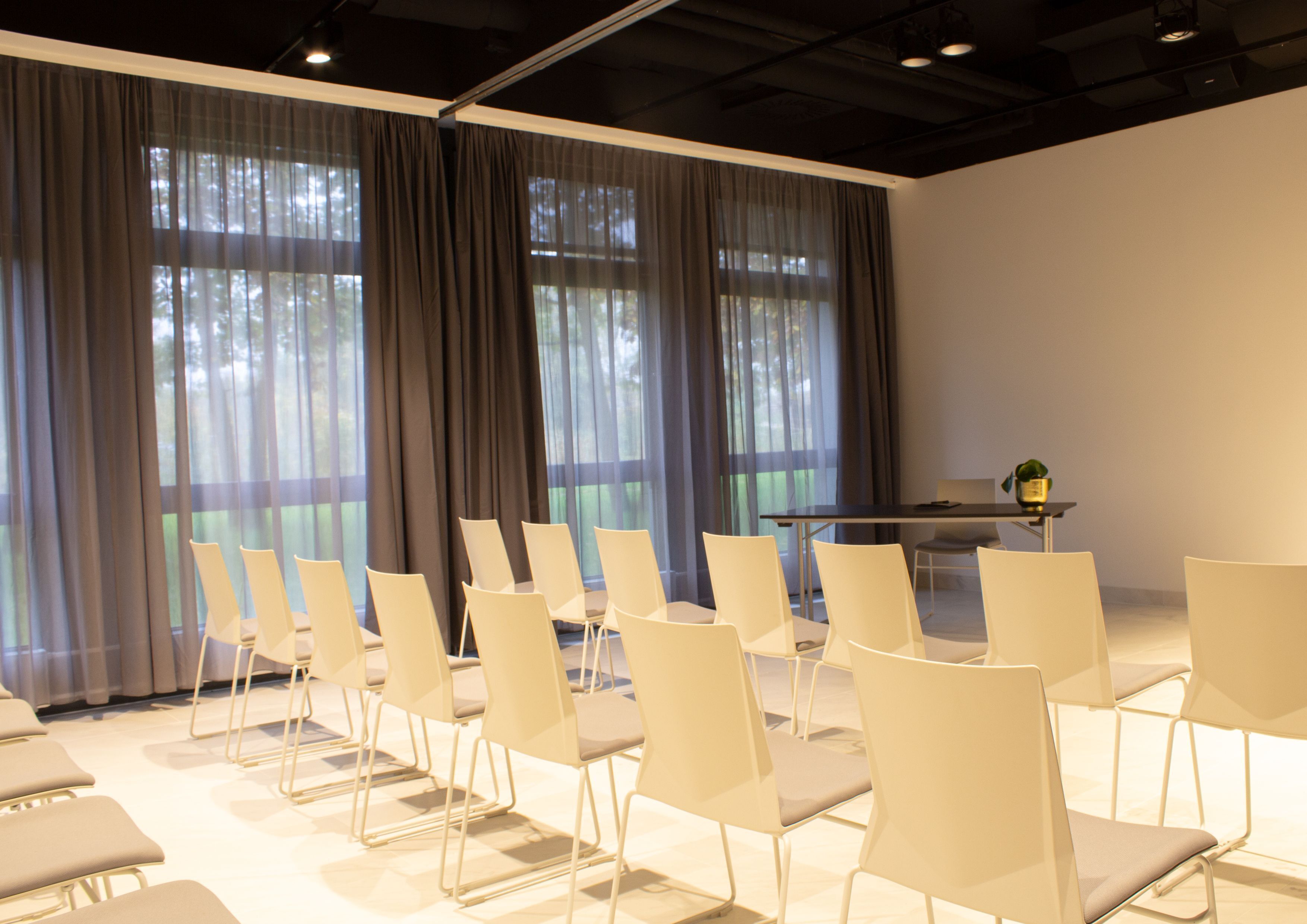 Un salón de conferencias con filas de sillas blancas, una mesa y ventanas amplias con cortinas translúcidas.