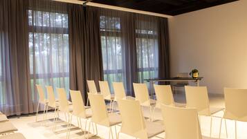 Un salón de conferencias con filas de sillas blancas, una mesa y ventanas amplias con cortinas translúcidas.