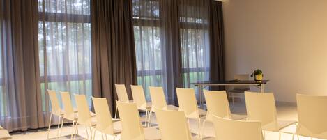 A conference room with rows of white chairs, a table, and large windows with sheer curtains.