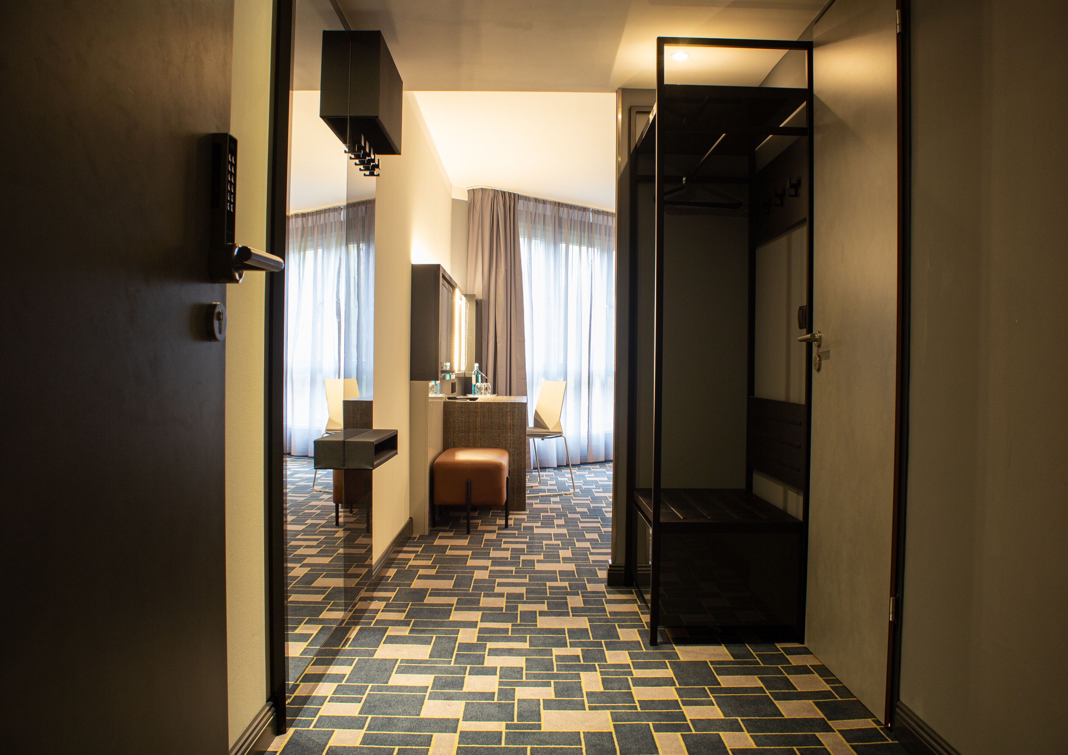 Un pasillo de hotel con piso estampado, un área de descanso con una silla y una mesita, y un espejo en la pared.