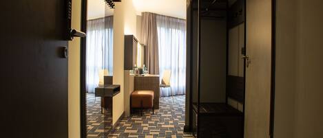 Un pasillo de hotel con piso estampado, un área de descanso con una silla y una mesita, y un espejo en la pared.