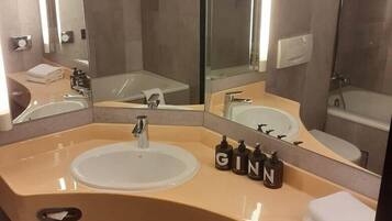 A modern bathroom with a curved beige countertop, a white sink, and a large mirror. There are two bottles labeled "GINN" on the countertop.
