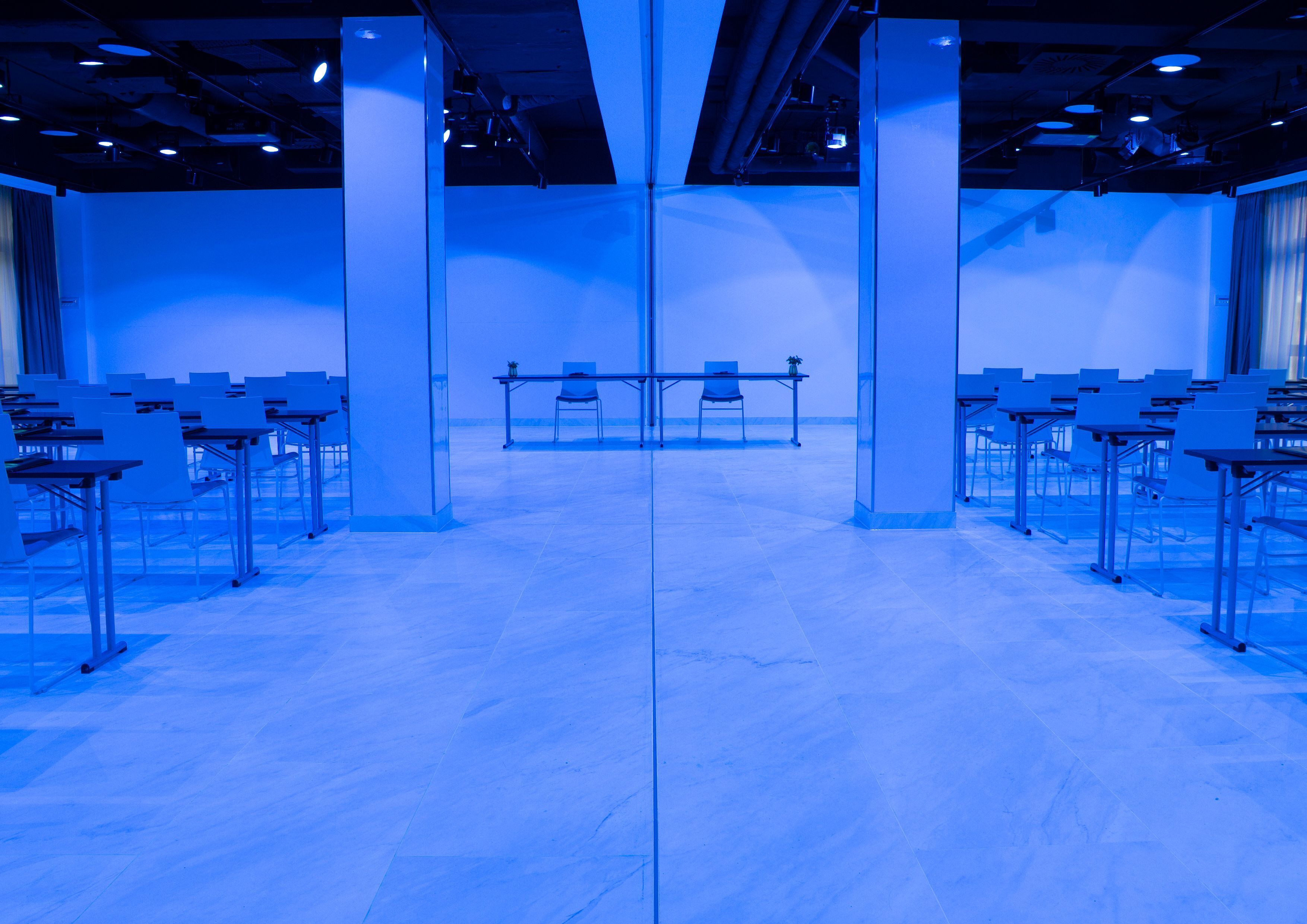A conference room with rows of tables and chairs, a stage at the front, and a large window on one side.