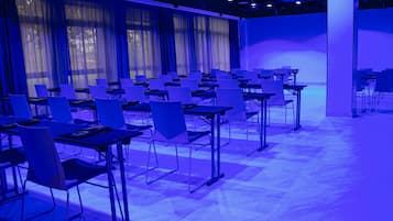 Uma sala de conferências com mesas e cadeiras dispostas em fileiras, grandes janelas com cortinas e teto com iluminação azul.