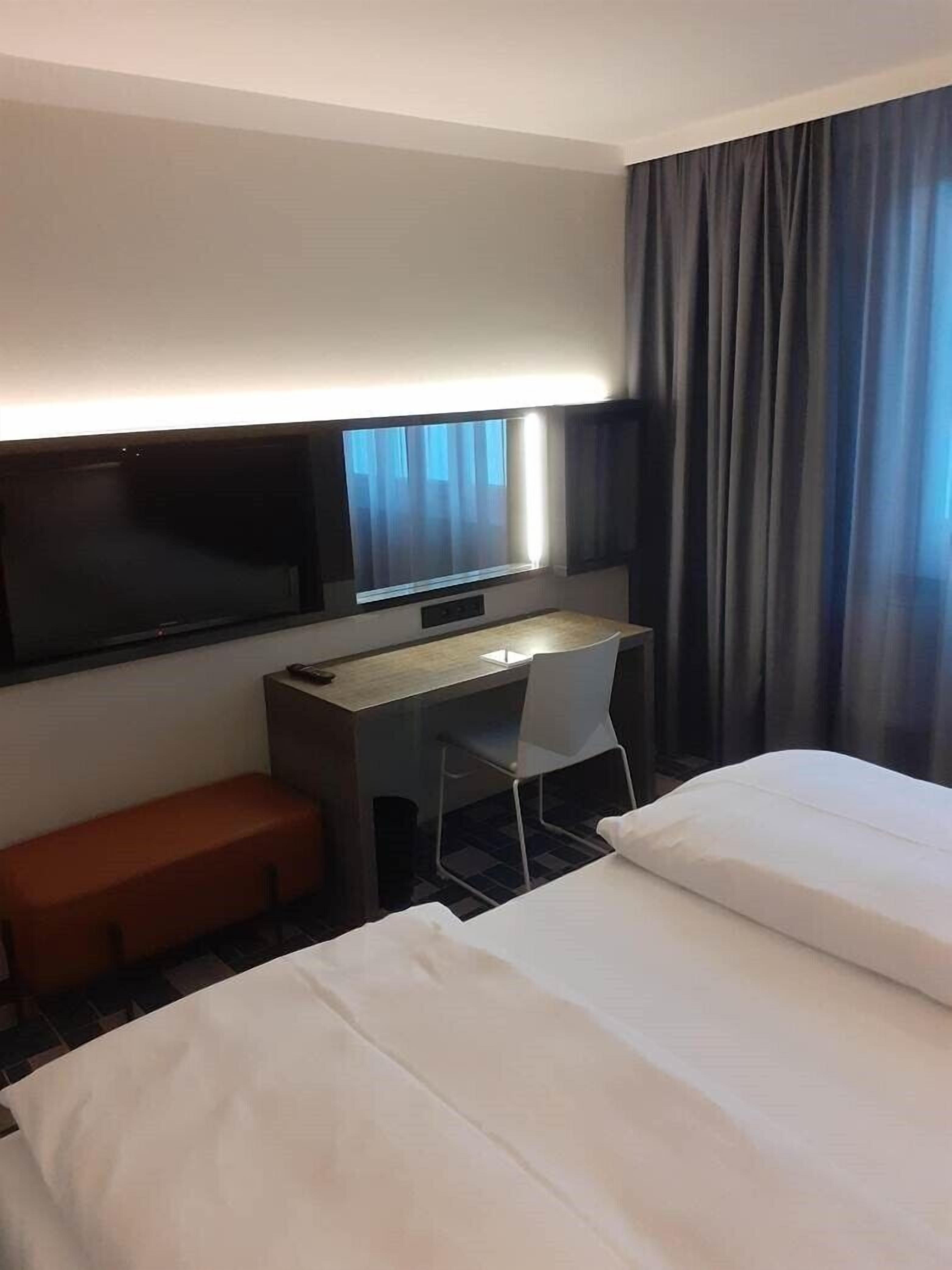A hotel room with a bed, a desk, a chair, a TV, and a window with curtains.