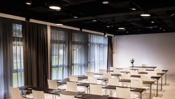 Un salón de conferencias con filas de mesas y sillas, amplias ventanas con cortinas, y un techo moderno con luminarias.