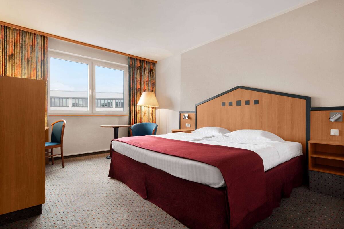 A hotel room with a large bed, a desk with a chair, a lamp, and a view of the outside.